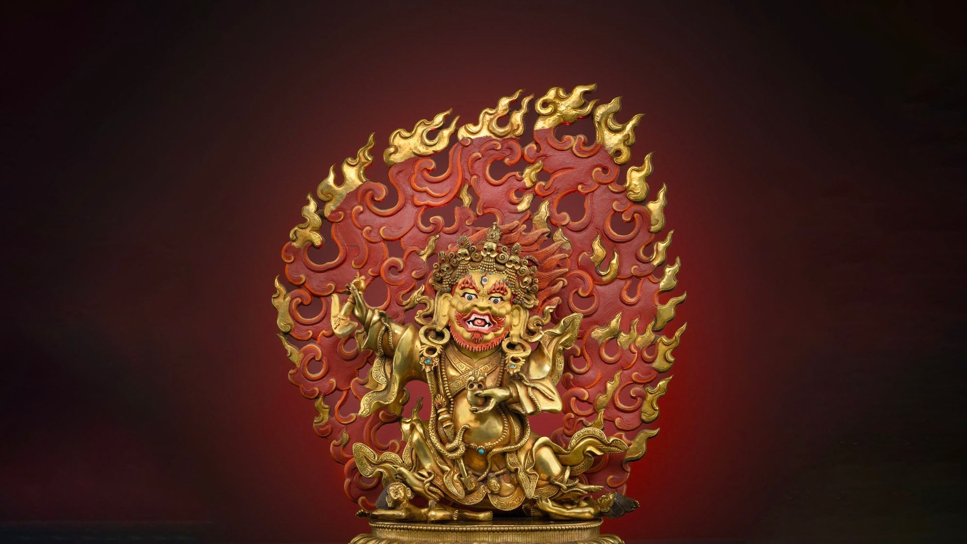 golden statue of Mahakala in a fierce position with red fiery backdrop with golden accents on the tip of the fire
