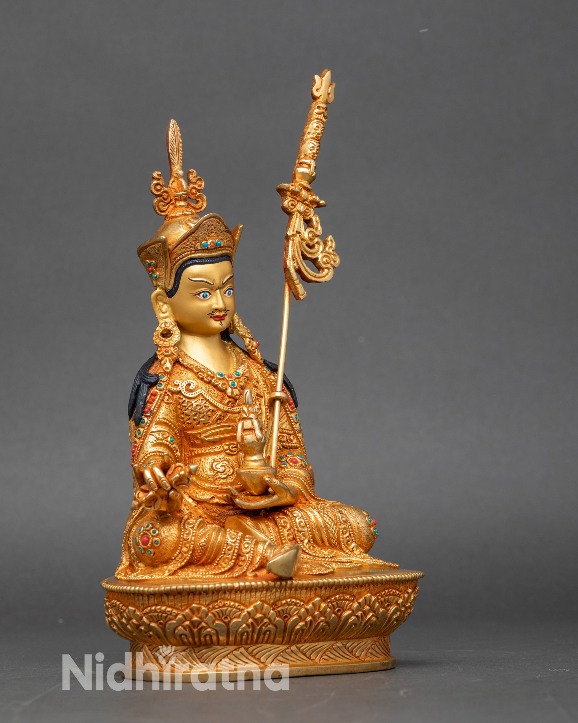 Side view of Guru Rinpoche statue with khatvanga resting on shoulder, gold-gilded artwork