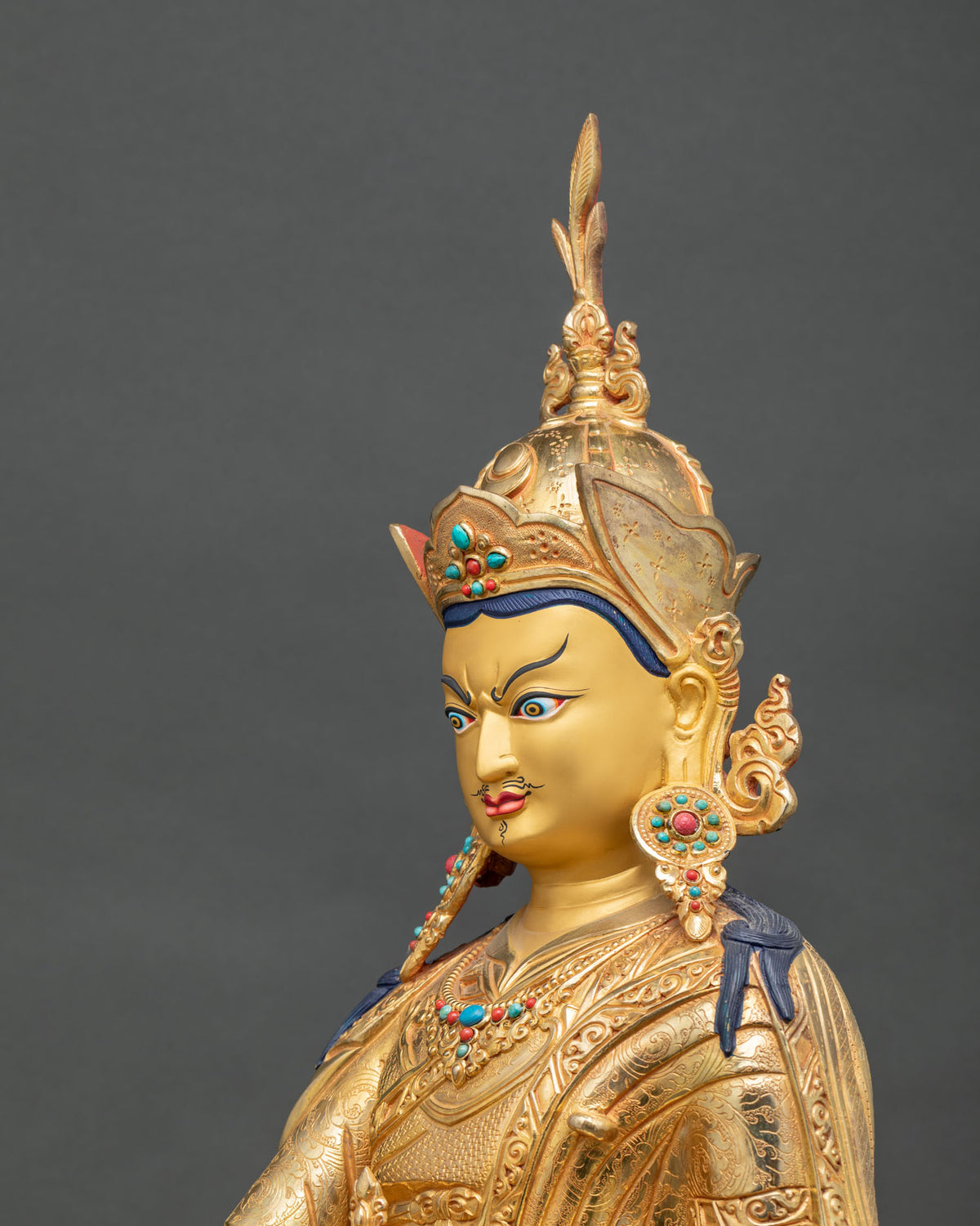 Angled close-up of the Padmasambhava face, lotus hat and gemstone earrings on a gold gilded statue