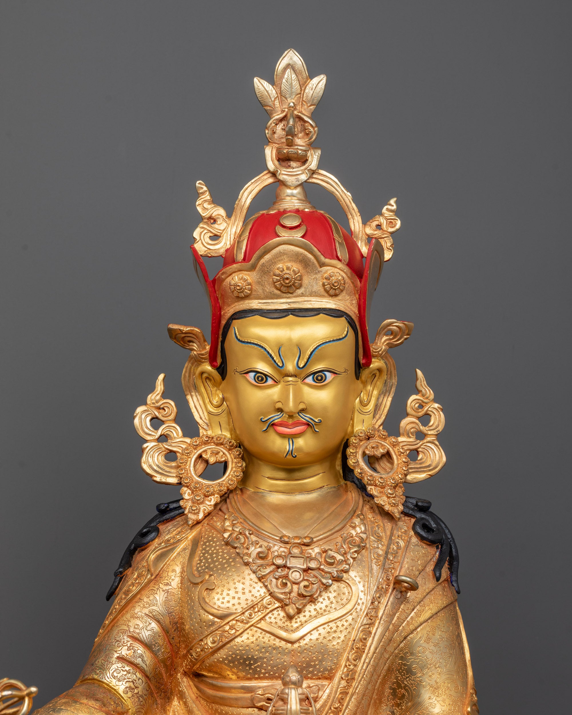 close up of Guru Rinpoche’s serene face and intricate jewelry on gold and fire-gilded body