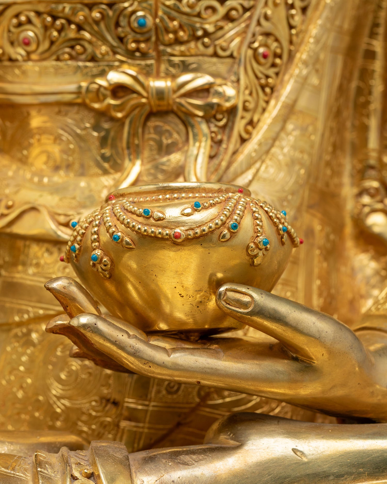 Close-up of turquoise and coral inlaid alms bowl held by gold Buddha statue
