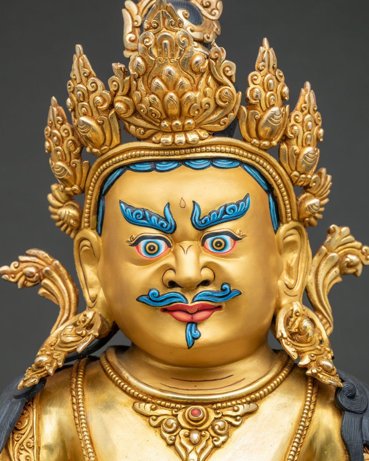 Detailed facial close up of gold gilded Dzambhala statue showing slightly wrathful expression.