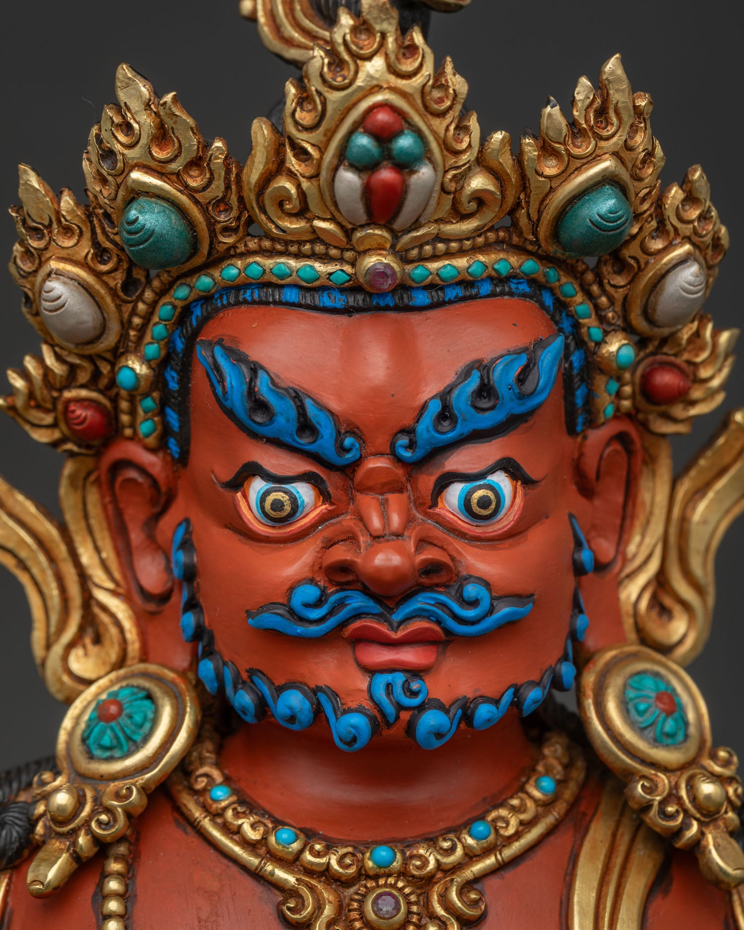 Face close up of Dzambhala statue highlighting blue beard, wrathful eyes, and turquoise five-jewel crown.