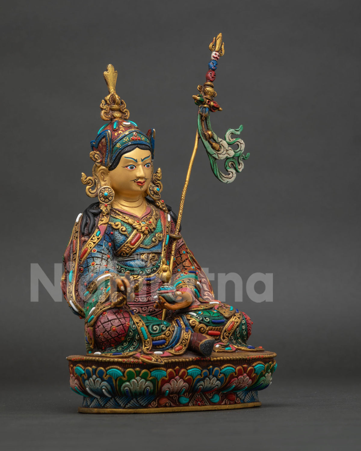 Guru rinpoche statue seated in royal ease on lotus throne holding vajra and skull cup.