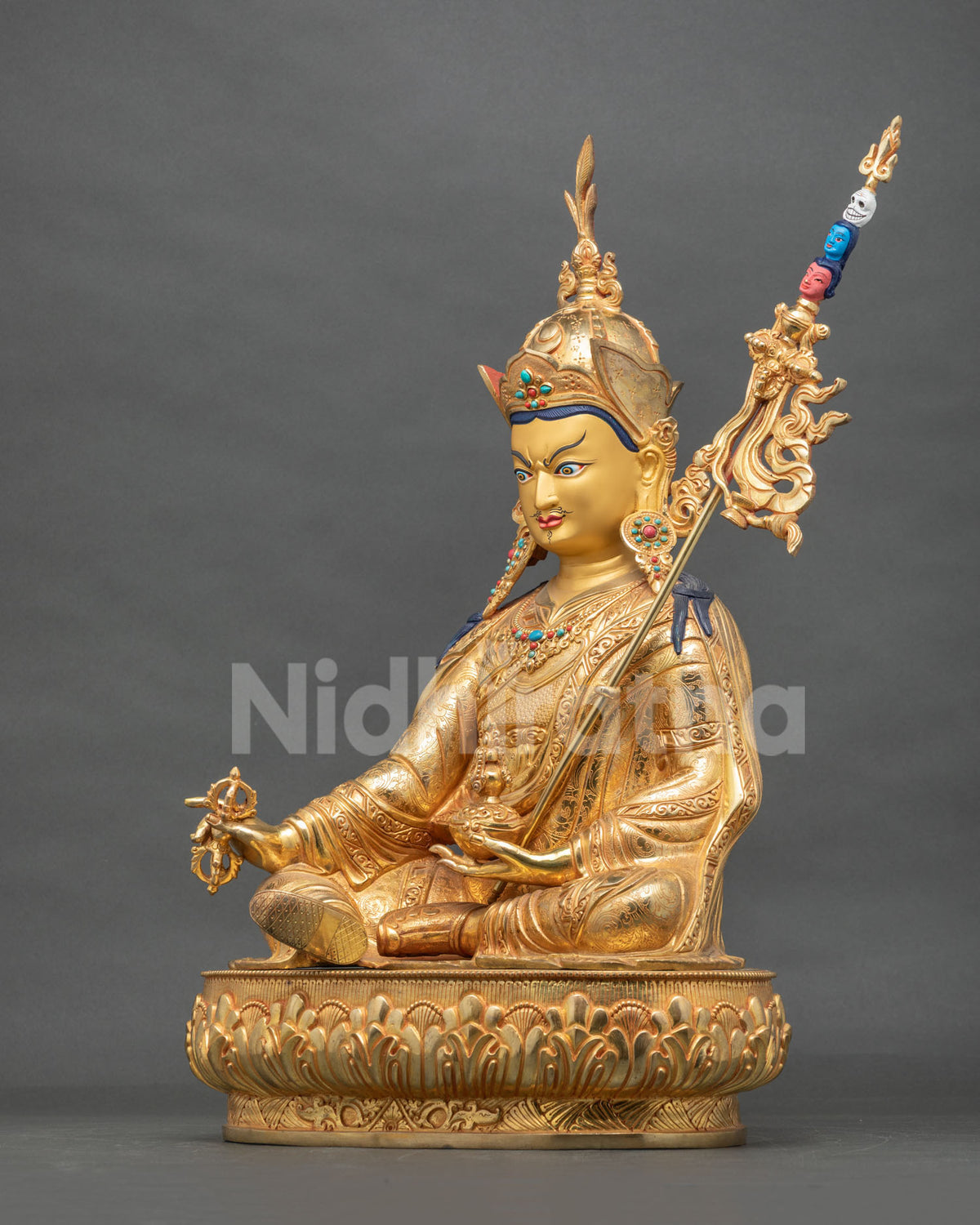Side view of the seated Guru Rinpoche copper statue with vajra and khatvanga staff on a lotus pedestal