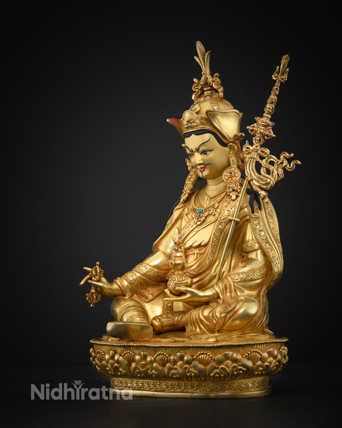Three-quarter view of gold-gilded Guru Rinpoche statue on an ornate lotus base, dark backdrop